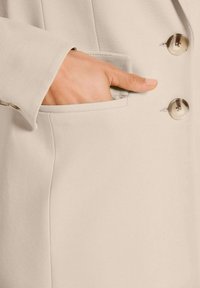 Hand met verzorgde nagels gedeeltelijk in de zak van een beige blazer, met grote bijpassende knopen zichtbaar.