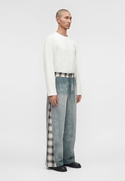 Άνδρας που στέκεται, φοράει ένα λευκό πουκάμισο με μακριά μανίκια και φαρδιά γκρι παντελόνια με καρό πλευρικές λωρίδες και μαύρα παπούτσια σε απλό φόντο.