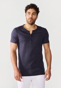 Camiseta de manga corta a rayas azul marino con cuello redondo y tapeta de dos botones. Tejido suave y textura lisa. Pantalones blancos visibles.