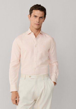 Hombre con camisa de botones de rayas rosa claro metida en pantalones plisados de color crema, de pie contra un fondo gris liso.
