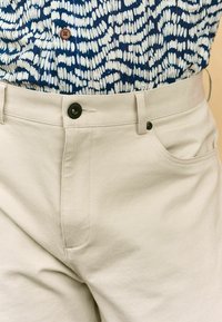 Pantalons en coton beige avec une finition texturée, dotés d'une fermeture à bouton à l'avant et de poches latérales, portés avec une chemise bleue à motifs.