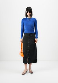 Vrouw met zwarte zonnebril, blauwe lange mouw top, zwarte knoop midi rok, zwarte open hakken, en een kleine oranje handtas.