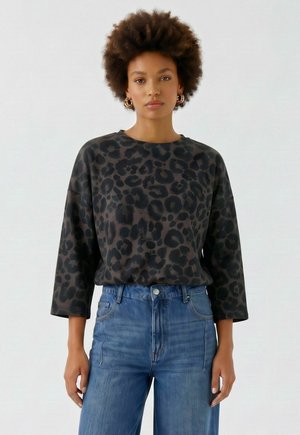 Vrouw met kort krullend haar draagt gouden creolen, een donker lang mouwen top met luipaardprint en een hoge blauwe spijkerbroek tegen een effen achtergrond.