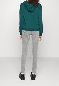 Sudadera verde con capucha ajustable y puños acanalados, combinada con jeans slim de color gris claro y zapatos negros de cordones, vista desde atrás.