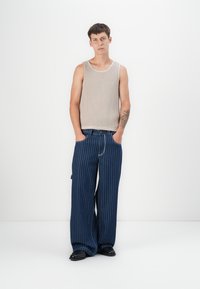 Top de tirantes acanalado en beige, pantalones holgados con rayas azul oscuro, combinados con zapatos negros. La modelo está de pie contra un fondo blanco liso.
