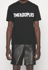 Svart bomullströja med fet vit text "THE KOOPLES." Kortsleeves med klassisk passform, i kombination med glänsande svarta lädershorts.