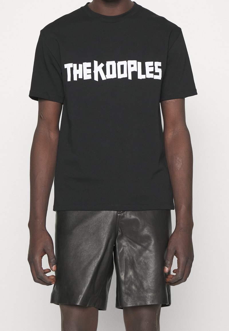 Svart bomullströja med fet vit text "THE KOOPLES." Kortsleeves med klassisk passform, i kombination med glänsande svarta lädershorts.