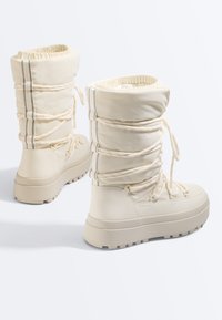 Bottes d'hiver crème isolées avec semelles épaisses, lacets à l'avant et bandes verticales réfléchissantes sur la tige matelassée.