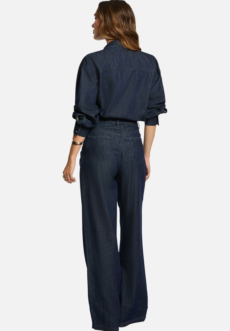 Chemise en denim foncé et jeans évasés, avec une taille haute, des manches retroussées et des poches arrière. Texture lisse avec des détails de coutures subtils.