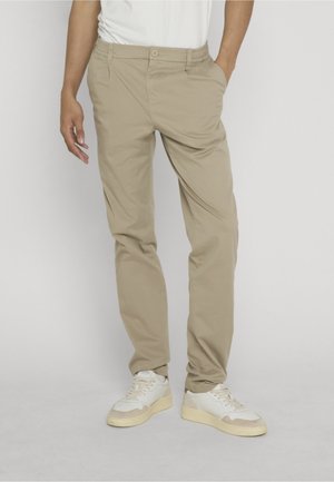 Mann iført beige chinos og hvite joggesko, stående med en hånd i lomma mot ensfarget bakgrunn.