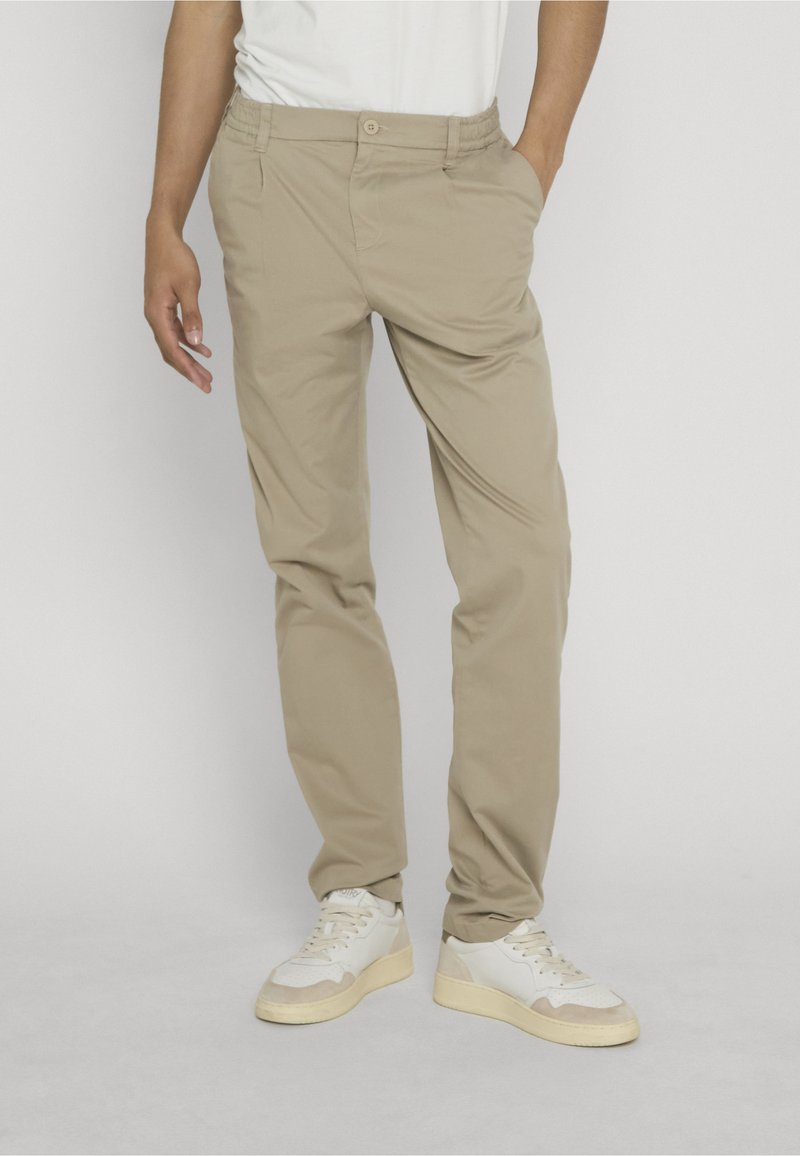 Homme portant un chino beige et des baskets blanches, debout une main dans la poche devant un fond uni.