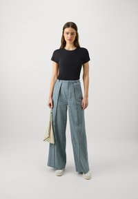 T-shirt nera attillata abbinata a pantaloni larghi azzurri chiari con pieghe; tiene in mano una borsa beige; ai piedi indossa sneakers bianche.