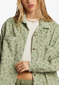 Camisa de pana verde con estampado de leopardo, con dos bolsillos frontales y botones marrones. Llevada sobre una camiseta color crema claro.