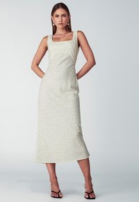 Cremefarbiges Midi-Kleid mit quadratischem Ausschnitt, schwarzen Pünktchen und tailliertem Oberteil. Weicher Stoff und seitliche Taschen. Getragen mit schwarzen Sandalen.