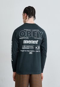 Long-sleeve black t-shirt featuring large white text on the back: "OBEY," "movement," and other logos; worn with plaid brown trousers.