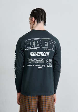 Schwarzes Langarm-T-Shirt mit großen weißen Texten auf dem Rücken: "OBEY", "movement" und anderen Logos; getragen mit karierten, braunen Hosen.