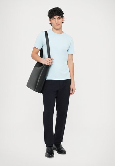 Μαύρη δερμάτινη τσάντα ώμου με μακριά ράβδο, που φοριέται από ένα άτομο με ανοιχτό μπλε μπλουζάκι και σκουρόχρωμο παντελόνι, στέκεται σε λευκό φόντο.