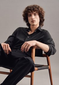 Camicia nera con bottoni, dalla texture liscia e colletto, abbinata a pantaloni neri sartoriali. Modello seduto su una sedia di legno con le braccia incrociate.