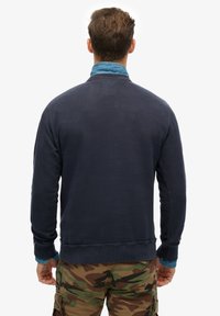 Marineblauwe sweatshirt met lange mouwen, geribbelde manchetten en een losse pasvorm, gecombineerd met een denim overhemd. Achtergrond is effen wit.
