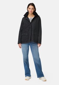 Zwarte waterdichte jas met een kraag, twee voorzakken en een knoopsluiting, gedragen over een gestreept shirt en gecombineerd met blauwe jeans.