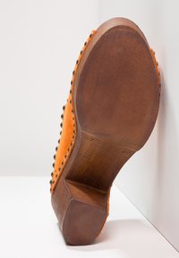 Chaussure orange en daim avec semelle en bois et clous en bronze le long du bord, talon relevé, photographiée de dessous sur fond blanc.