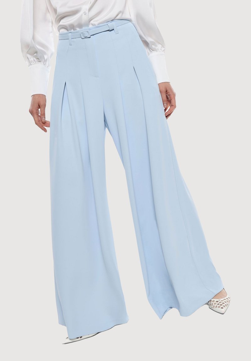 Pantaloni a gamba larga blu chiaro con vita cinturata, pieghe sul davanti e una texture del tessuto liscia, abbinati a una blusa bianca e tacchi.