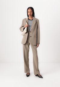 Beige skreddersydd dress med enkelt kneppet blazer, stripete topp under, og svarte spisse leiligheter. Holder en liten beige håndveske.
