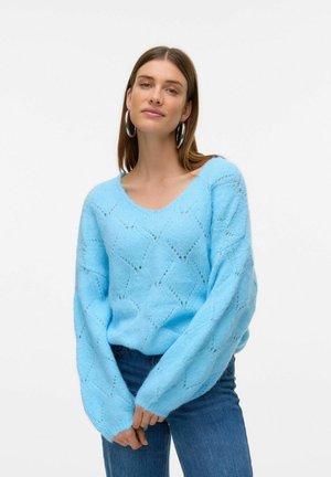 Himmelblauer Strickpullover mit V-Ausschnitt, der mit Rautenmuster und lockeren Ärmeln versehen ist. Getragen mit dunklen Jeans vor einem weißen Hintergrund.