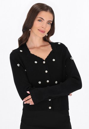 Vrouw met lang bruin haar draagt een zwarte cardigan versierd met parelknopen en parel bloemmotieven, armen gekruist, licht glimlachend.