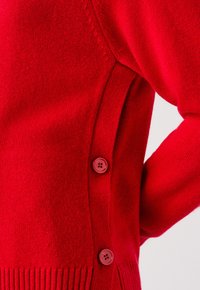 Czerwony sweter z dzianiny z prążkowanymi krawędziami, z trzema pasującymi czerwonymi guzikami po boku i gładką fakturą. Zbliżenie na materiał.