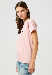 T-shirt in cotone rosa con maniche corte e scollatura rotonda, caratterizzato da un piccolo logo blu sulla parte superiore del petto. Texture liscia.