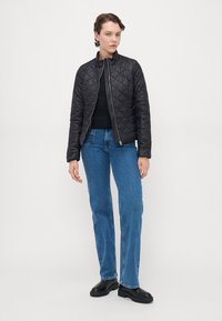 Svart quiltet jakke med høy krage, kombinert med en svart topp og blå rettleggete jeans. Har glidelås foran og et minimalistisk design.