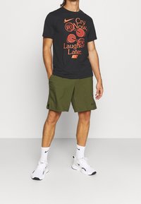 Camiseta negra de Nike con el texto gráfico en naranja "Llora ahora, ríe después"; pantalones cortos verde oliva; zapatillas blancas con acentos negros; calcetines blancos.