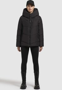 Giacca a vento nera con cappuccio, dotata di chiusura con zip, cuciture a zig zag e tasche con zip frontali. Abbinata a leggings neri e stivali da combattimento.