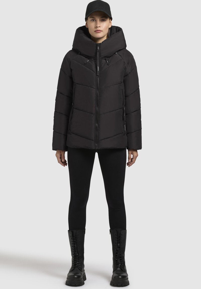 Giacca a vento nera con cappuccio, dotata di chiusura con zip, cuciture a zig zag e tasche con zip frontali. Abbinata a leggings neri e stivali da combattimento.