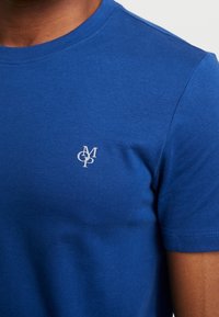 Camiseta de algodón azul con cuello redondo y mangas cortas. Presenta un pequeño logo blanco en el pecho. Textura suave.