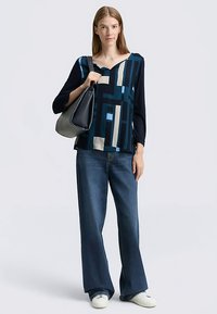 Blue and black patterned blouse with a V-neck, paired with wide-leg blue jeans and white sneakers, holding a gray handbag.