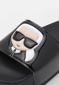 Sandale noire à bride brillante avec un visage stylisé portant des lunettes de soleil noires et des détails blancs sur les cheveux et le col.