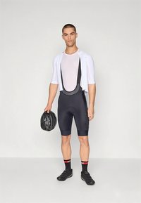 Černé cyklistické kalhoty s laclem a bílý síťovaný top, drží černou helmu. Černé ponožky s červenými detaily a černé cyklistické boty.