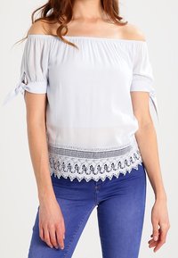 Top azzurro chiaro scollato sulle spalle con maniche corte e dettagli annodati. Presenta un bordo in pizzo nella parte inferiore e un tessuto testurizzato. Design casual.