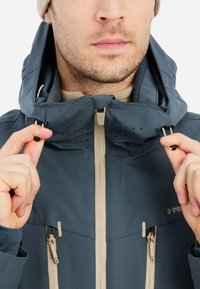 Wasserabweisende blaue Jacke mit hohem Kragen, beigen Akzenten, verstellbaren Kordeln und Belüftungslöchern in der Nähe der Kapuze.