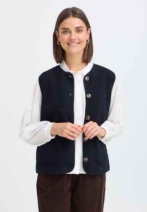 Jeune femme aux cheveux courts bruns portant un chemisier blanc, un gilet polaire bleu marine boutonné et un pantalon marron, souriante avec les mains près des boutons.