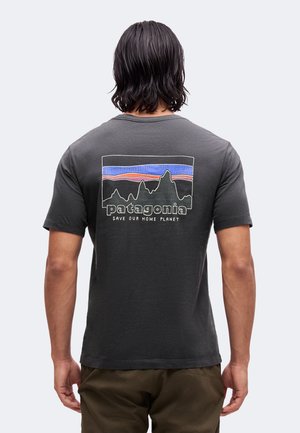 Person med skulderlangt mørkt hår iført en mørkegrå Patagonia t-shirt med en farverig bjerglogo og teksten "Save Our Home Planet" på ryggen.