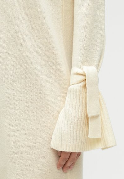 Μπεζ πλεκτό πουλόβερ με φαρδιές ριγέ μανσέτες δεμένες με κορδέλα από το ίδιο ύφασμα. Η υφή είναι μαλακή και ελαφρώς χαλαρή πλέξη.