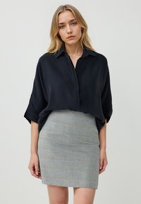 Chemisier noir ample avec un col ; associé à une jupe ajustée grise à carreaux comportant de petits motifs et une texture lisse.