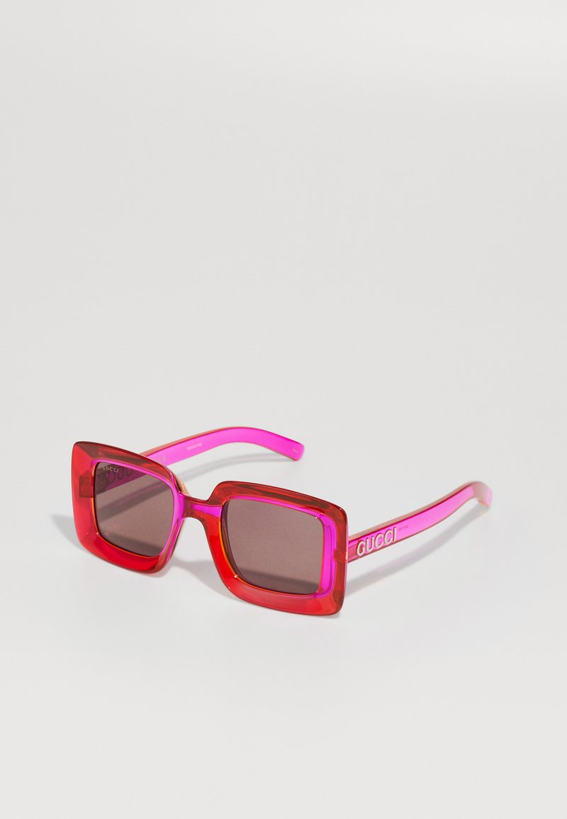 Røde og rosa firkantede solbriller med brune linser, som har en gjennomsiktig ramme og en logo på stangene. Glatt og shiny overflate.