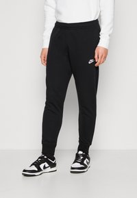 Persona vistiendo joggers negros de Nike y zapatillas negras y blancas de Nike, de pie contra un fondo gris claro liso.