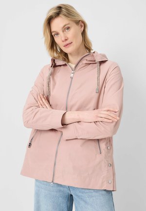 Blonde woman with blue eyes wearing a light pink zip-up jacket with hood and drawstrings, paired with light blue jeans, arms crossed.