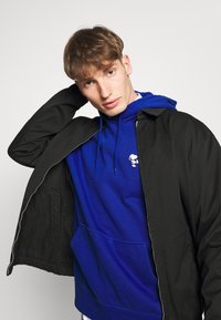 Giacca nera con zip sopra una felpa blu, dotata di una tasca frontale e di un piccolo logo di Snoopy. Tessuto morbido, design casual.