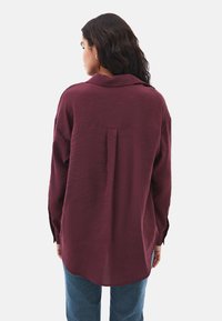 Camicia button-up bordeaux con colletto, vestibilità rilassata, dettaglio con piega sul retro e maniche lunghe, abbinata a jeans blu.